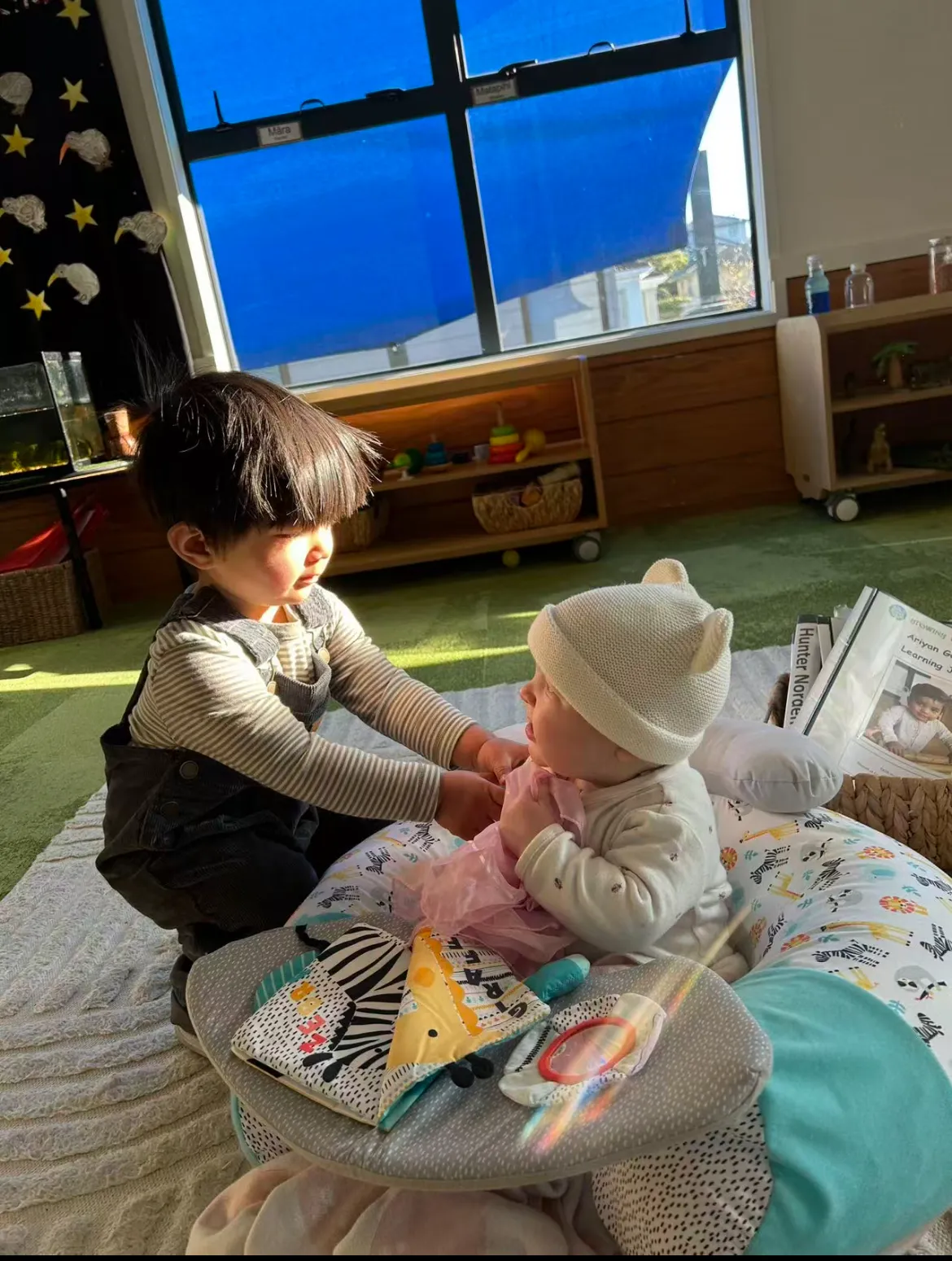 A toddler gently reaching out to a baby resting on a support pillow in the Fantail Room at Growing Minds